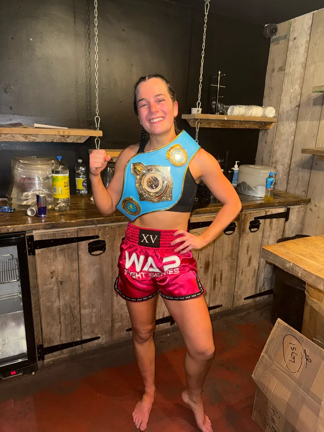 A fighter in pink WAR Fight Series shorts wearing a championship belt over one shoulder in a backstage room