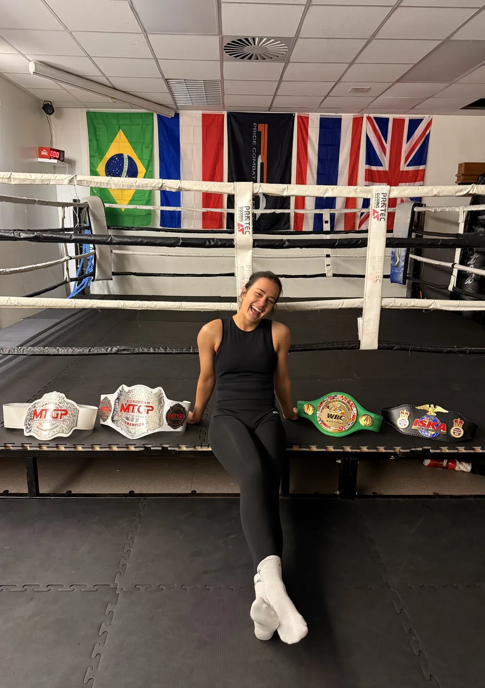 Five championship belts lined up along the apron of a boxing ring in a gym, under national flags on the wall